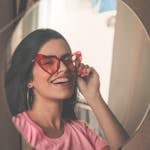 Stylish woman wearing heart-shaped sunglasses smiles joyfully at her reflection in a mirror.